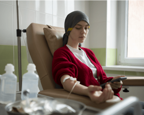 A young woman sitting in a medical chair, wearing a red cardigan and a headscarf, while receiving treatment. She is holding a smartphone and has an IV drip connected to her arm.