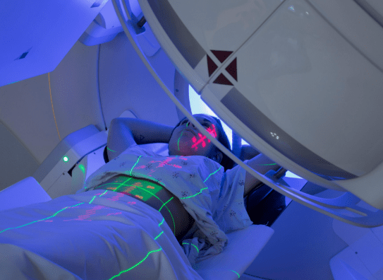 A patient lying on a treatment bed inside a medical machine, with laser markings on their body and blue illumination in the environment.
