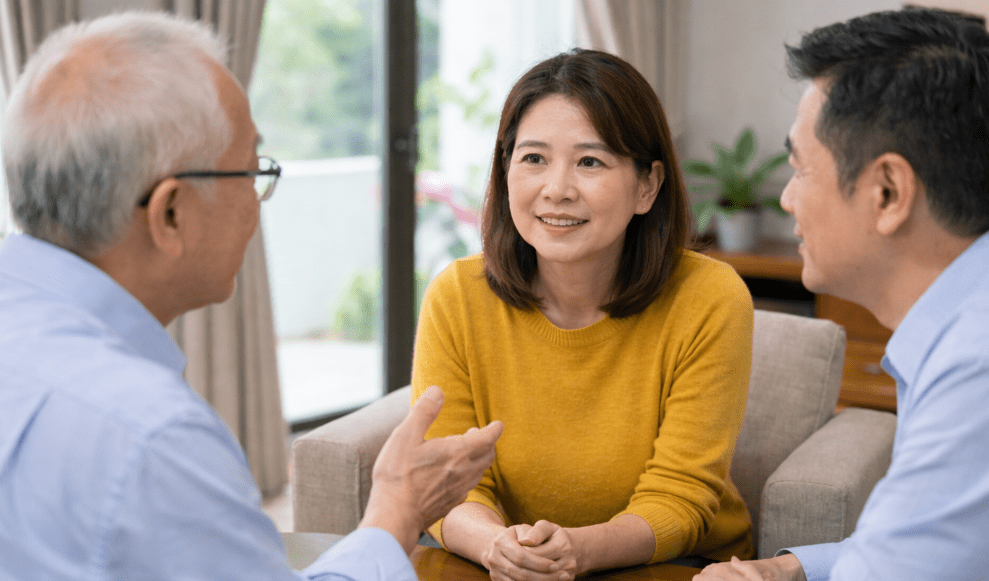 A woman in a yellow sweater engages in conversation with two men, one older and one younger, in a bright, indoor setting.
