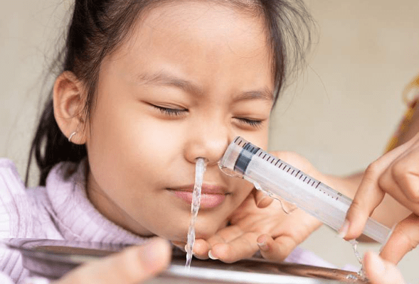 A young girl closes her eyes as water is administered through a syringe into her nose, with an adult's hand assisting. A metal tray is placed underneath to catch the liquid.