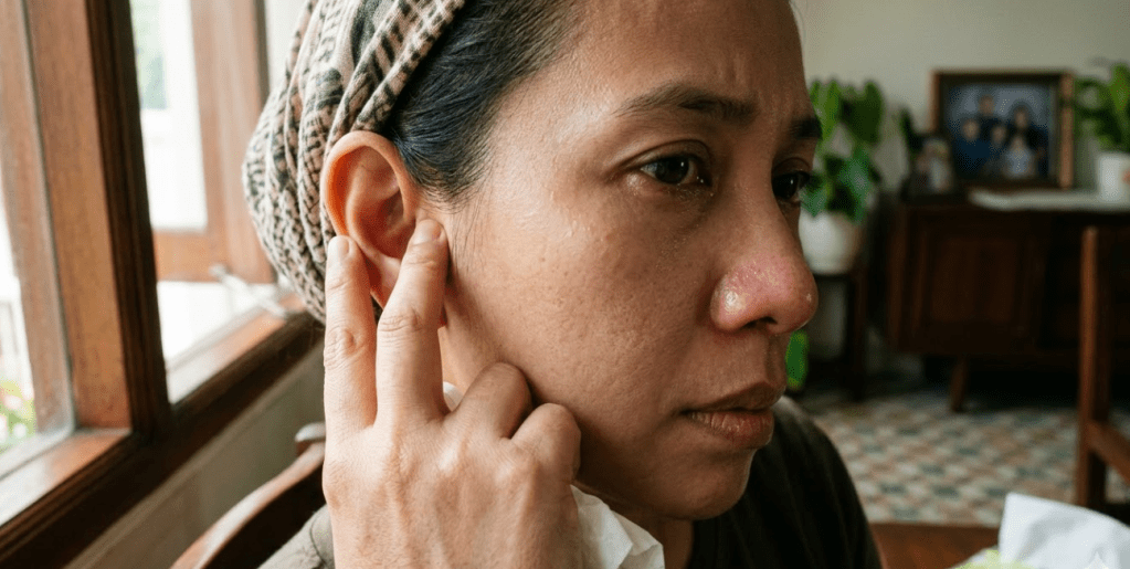 A woman with a headscarf sits indoors, touching her ear with a concerned expression, tears in her eyes, and a tissue in her other hand.